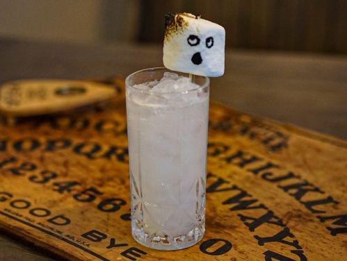 A clear drink with ice, topped with a toasted marshmallow featuring a ghostly face, on a wooden tray.