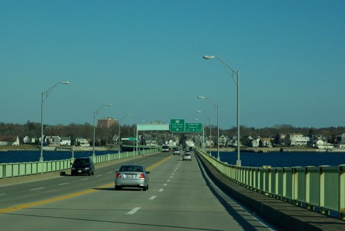 A view of a bridge with cars driving, leading to a green highway sign over a body of water and houses in the background.