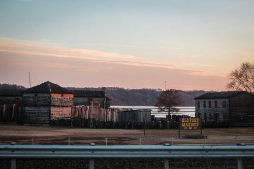 Historic wooden buildings at Old Fort Madison, with a river and trees in the background during sunset.