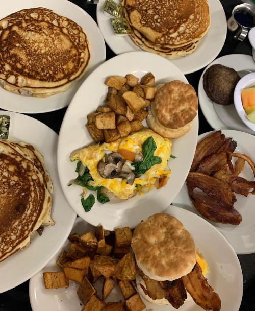 A spread of breakfast items including pancakes, eggs with spinach and mushrooms, bacon, potatoes, and fruit.