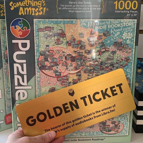 A hand holds a golden ticket in front of a colorful puzzle box featuring a Midwest indie bookstore roadmap.