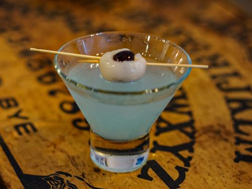 A clear cocktail in a glass, garnished with a lychee and a cherry on a skewer, resting on a wooden surface.