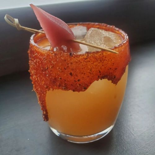 A colorful cocktail with a sugared rim, ice, and a slice of fruit on a skewer, set against a dark background.