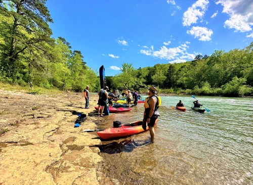 A group of people preparing kayaks by a river on a sunny day, surrounded by lush greenery and blue skies.