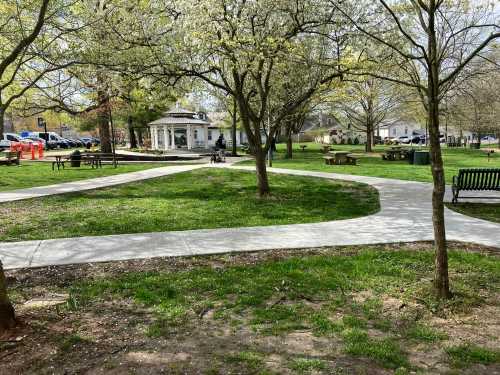 A park scene with winding paths, green grass, trees, and a gazebo in the background on a sunny day.