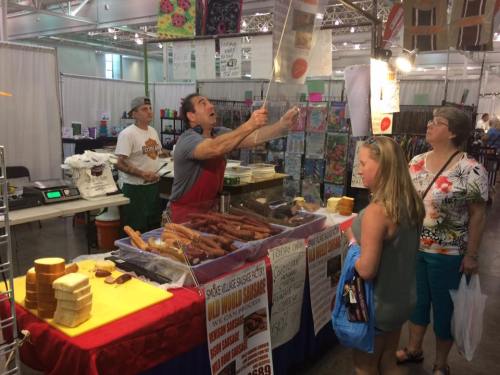 A vendor enthusiastically promotes sausages at a market, while customers browse nearby.