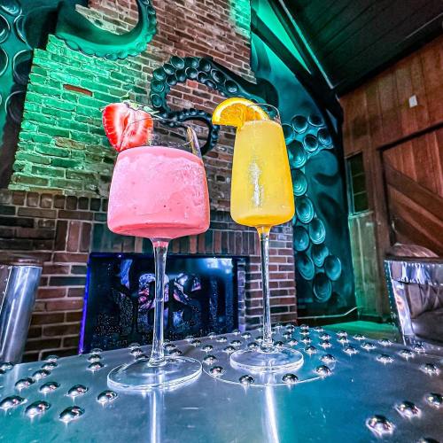Two colorful cocktails on a metallic table, one pink with a strawberry and one orange with an orange slice, against a brick wall.