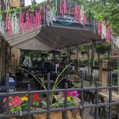 A cozy outdoor dining area with a tent, colorful flowers, and string lights, surrounded by greenery.