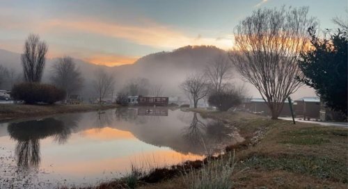 A serene landscape at dawn, featuring a misty pond, trees, and mountains under a colorful sky.