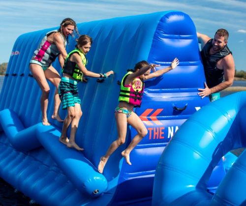 Four people, including two children, play on a large blue inflatable water slide, enjoying a sunny day by the water.
