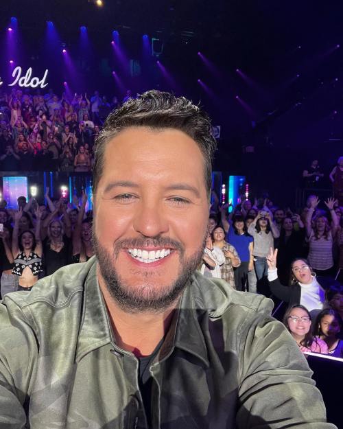 A smiling man takes a selfie with a cheering crowd in the background, illuminated by colorful stage lights.