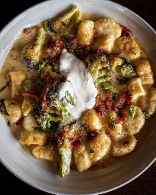 Creamy gnocchi dish topped with broccoli, bacon, and a dollop of sour cream, garnished with herbs.