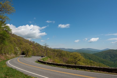 A winding road curves through lush green mountains under a clear blue sky with scattered clouds.