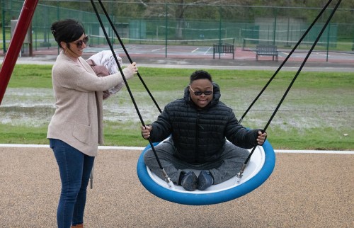 A child in sunglasses swings on a large swing while an adult holds a baby nearby at a playground.
