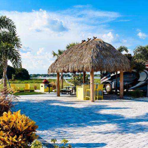 A tropical outdoor space with a thatched gazebo, yellow furniture, and lush greenery under a blue sky.
