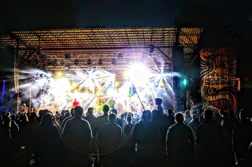 A crowd watches a vibrant concert at night, illuminated by bright stage lights and colorful decorations.