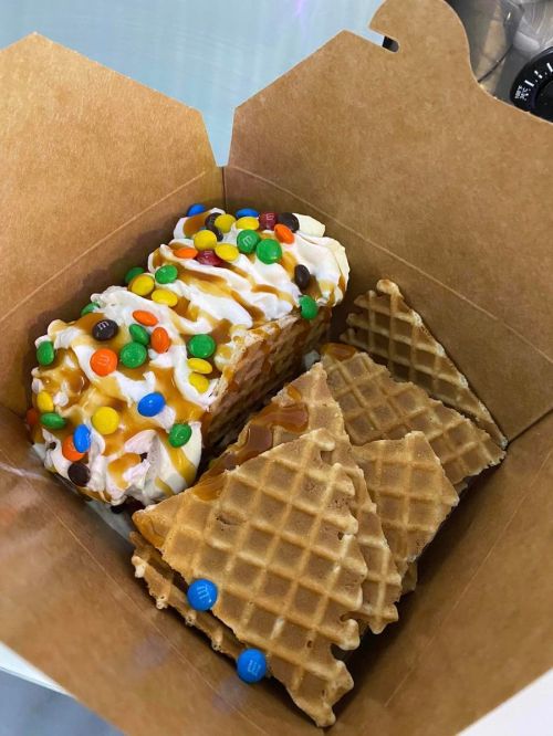 A takeout box containing a dessert with whipped cream, caramel, chocolate candies, and waffle pieces.