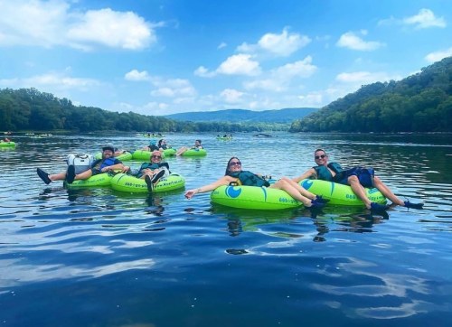 Group of people relaxing on green inner tubes in a calm river, surrounded by trees and blue sky.