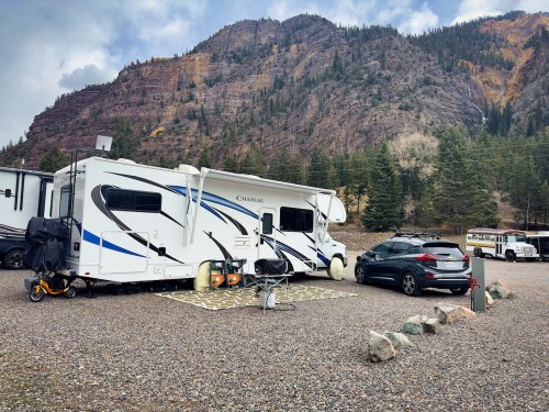 A parked RV next to a car in a gravel area, surrounded by mountains and trees.