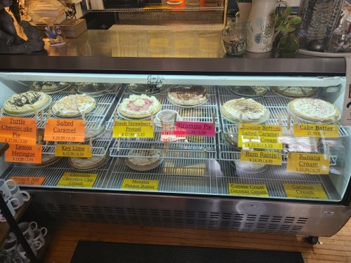 A display case filled with various pies, including turtle cheesecake, salted caramel, and fresh strawberry, among others.
