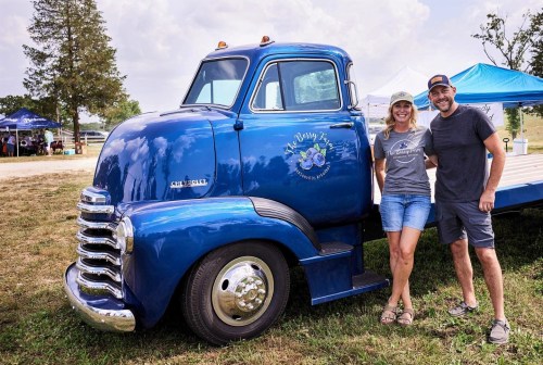 A couple stands beside a vintage blue Chevrolet truck at an outdoor event, with tents in the background.