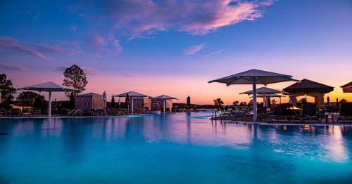 A serene pool at sunset, with lounge chairs and umbrellas, reflecting vibrant colors in the water.
