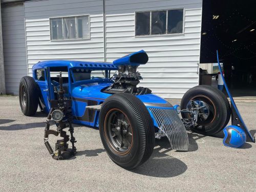 A custom blue hot rod with a large engine and unique design, parked beside a mechanical sculpture.