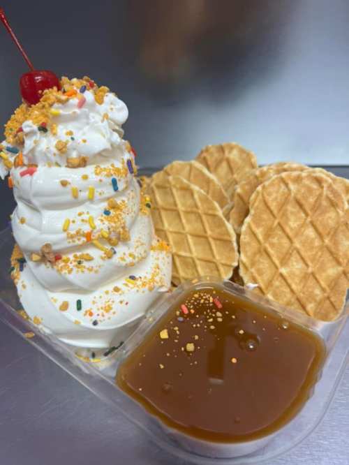 A swirl of soft-serve ice cream topped with sprinkles and a cherry, served with waffle cookies and caramel sauce.
