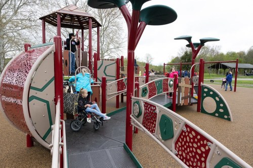 A colorful playground with a slide, ramps, and children playing, including one in a stroller. Trees in the background.