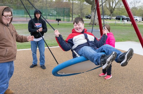 A boy swings joyfully on a playground swing, while friends assist and watch nearby on a cloudy day.