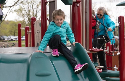 A young girl in a turquoise jacket and pink shoes climbs down a slide at a playground, smiling at the camera.