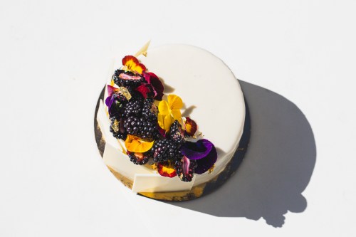 A round cake topped with blackberries and colorful edible flowers on a white background.