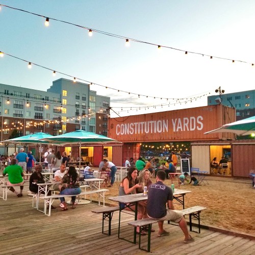 Outdoor dining area at Constitution Yards, featuring string lights, picnic tables, and people enjoying the evening.