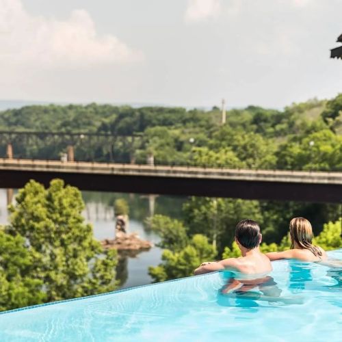 A couple relaxes in an infinity pool overlooking a river and lush greenery on a sunny day.