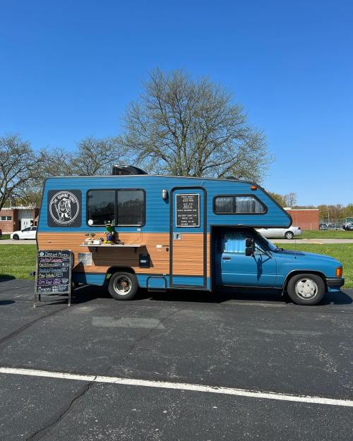 A colorful food truck parked on a sunny day, featuring a menu board and surrounded by green trees.