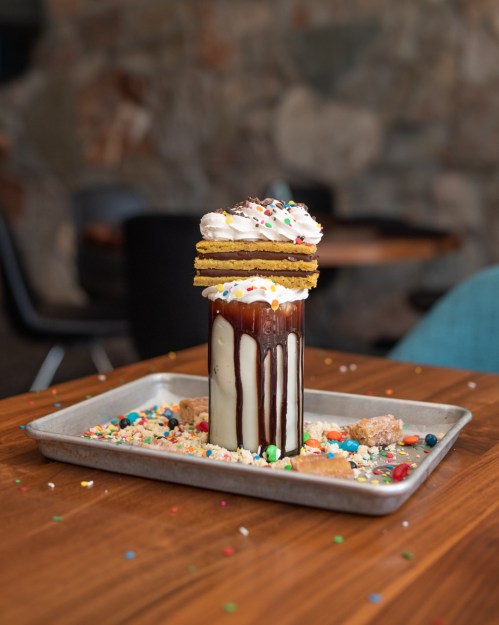 A tall dessert drink topped with whipped cream, cookies, and colorful sprinkles, served on a tray with scattered toppings.