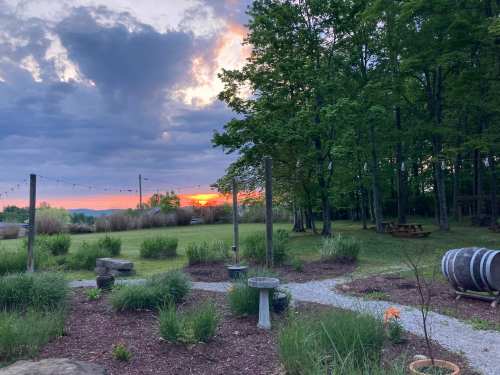 Sunset over a grassy area with trees, picnic tables, and garden features, under a cloudy sky.
