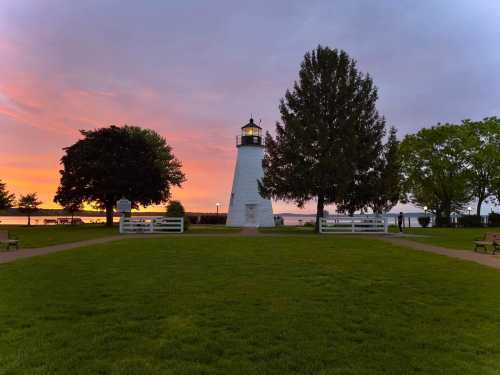 A white lighthouse stands in a park at sunset, surrounded by trees and benches, with a colorful sky in the background.