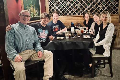 A group of six people sitting around a table in a restaurant, enjoying food and drinks, smiling at the camera.