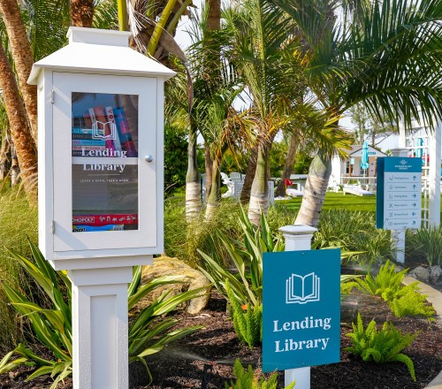 A white lending library box surrounded by palm trees and greenery, with a sign indicating "Lending Library."