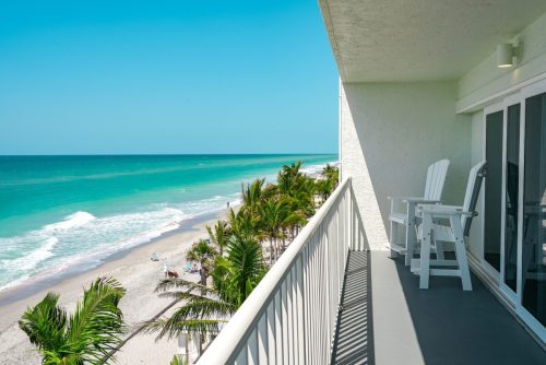 A balcony view overlooking a sandy beach with turquoise waves and palm trees under a clear blue sky.