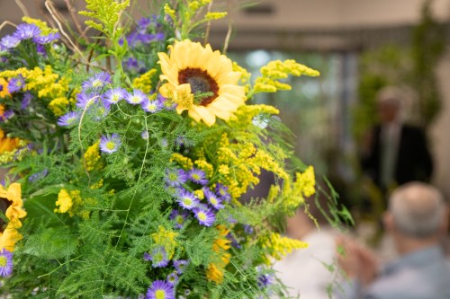 A vibrant floral arrangement featuring sunflowers and purple flowers, with a blurred background of people at an event.