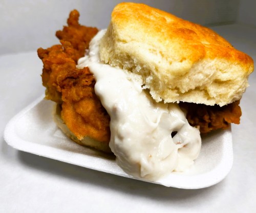 A close-up of a fried chicken biscuit sandwich with creamy gravy on a white paper tray.
