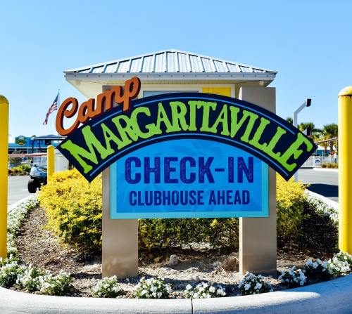 Colorful sign for Camp Margaritaville, indicating check-in for the clubhouse ahead, surrounded by flowers and blue sky.