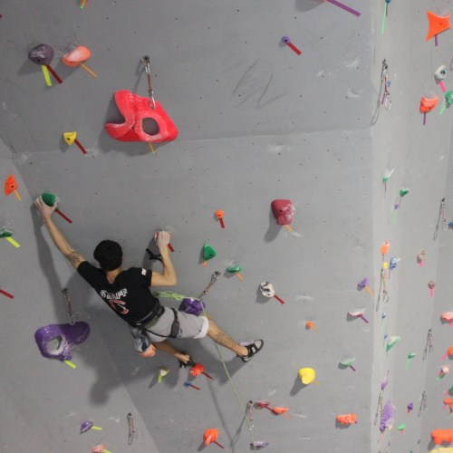 A climber ascends a colorful indoor rock wall, using various holds and wearing climbing gear.