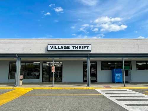 Exterior of a thrift store named "Village Thrift," featuring a blue donation bin and a clear sky above.