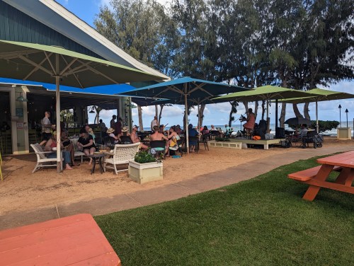 A lively outdoor dining area with people seated under colorful umbrellas, overlooking a beach and ocean.