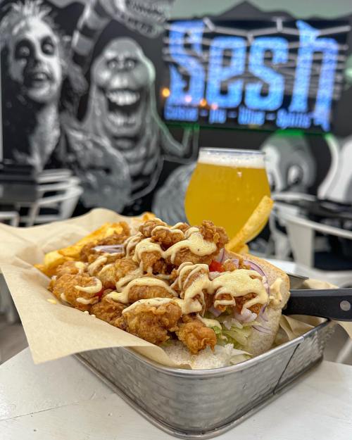 A basket of fried chicken with toppings and fries, alongside a glass of beer, set against a vibrant mural backdrop.
