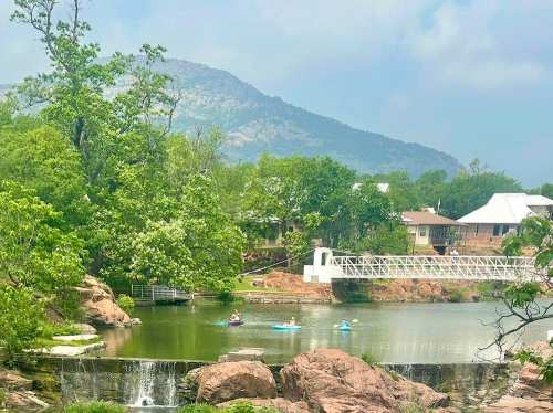 A serene lake surrounded by greenery, with kayakers paddling and a bridge in the background against a mountain backdrop.