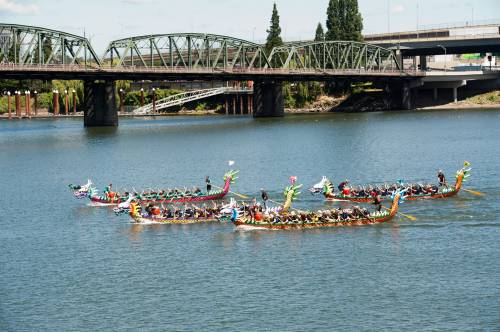 Colorful dragon boats race on a river, with a bridge and trees in the background under a clear blue sky.